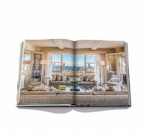 An open book, The Carlyle by Assouline, shows a modern living room with large windows overlooking a city skyline, styled with neutral sofas, a glass coffee table, and decorative lamps—evoking luxury hotel suite elegance.