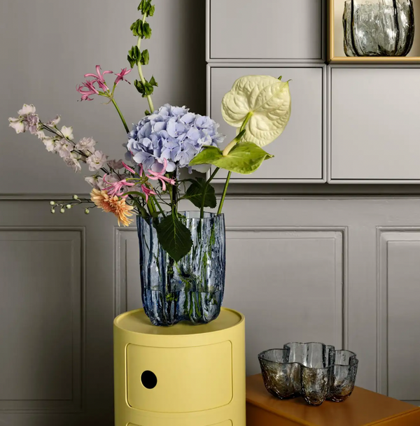 A bouquet featuring blue hydrangea, pink lilies, and orange gerberas is arranged in the Crackle Bowl Blue by Orrefors Kosta Boda, placed on a yellow side table.