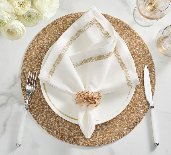 A formal tablescape featuring the Kim Seybert Impression White & Gold Napkin, a gold-patterned charger, white plate, silverware, two glasses, a decorative napkin ring, and white flowers on marble.