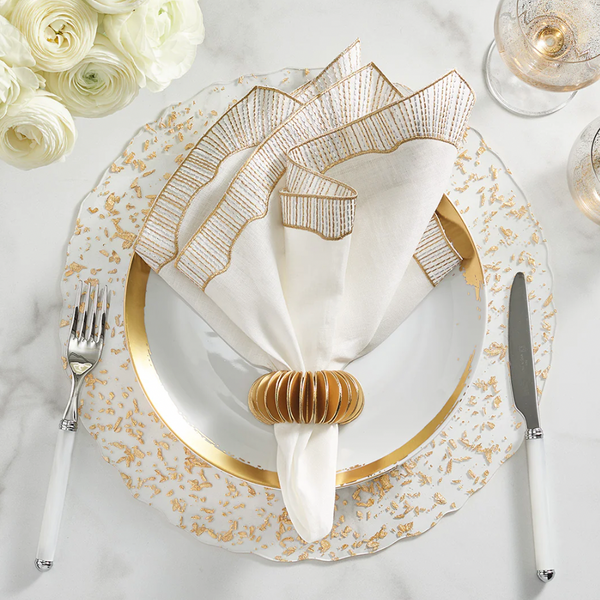 Elegant table setting featuring Kim Seybert's Frame White, Gold & Silver Napkin Set of 4, paired with a gold napkin ring, white and gold-rimmed plate, white-handled flatware, two glasses, and white flowers on a marble surface.