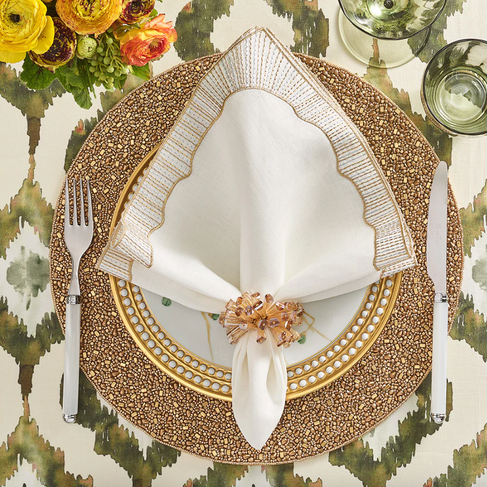 A formal place setting with a gold and white plate, beaded placemat, Kim Seybert Frame White, Gold & Silver Napkin set held by a ring, flatware, green glasses, floral centerpiece, and patterned tablecloth.