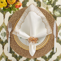 A formal place setting with a gold and white plate, beaded placemat, Kim Seybert Frame White, Gold & Silver Napkin set held by a ring, flatware, green glasses, floral centerpiece, and patterned tablecloth.