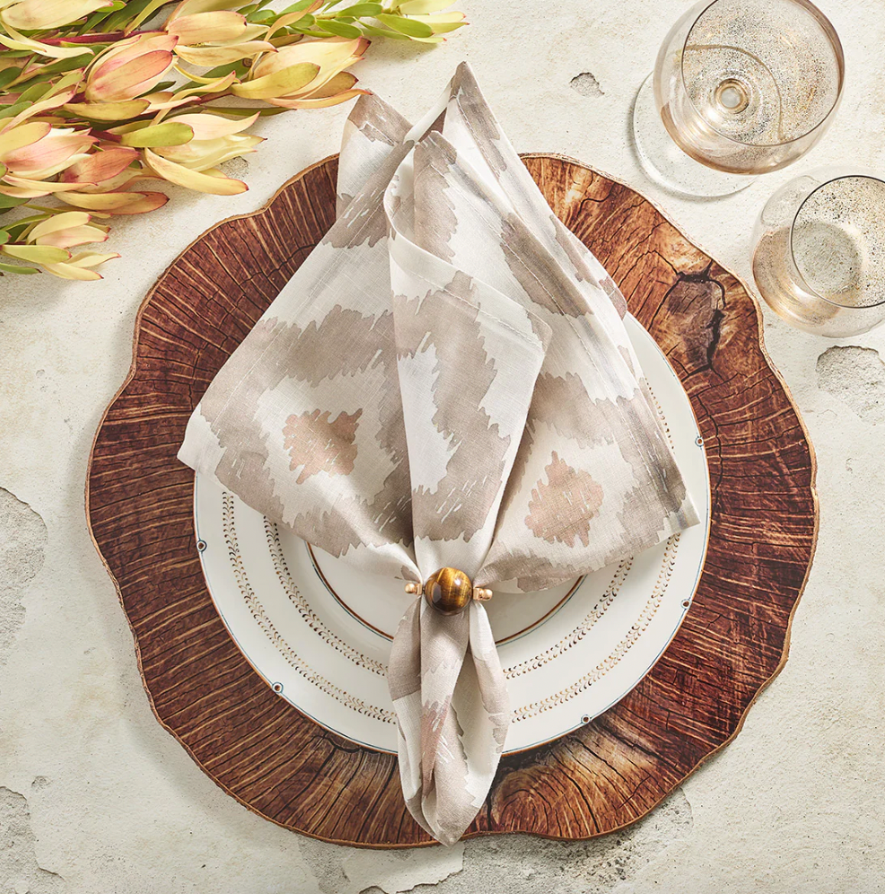 A decorative setting features layered plates, a wood-textured charger, Kim Seybert’s Watercolor Ikat Napkin Taupe (set of 4) folded in a fan with a wooden ring, two clear glasses, and light yellow flowers on a textured surface.