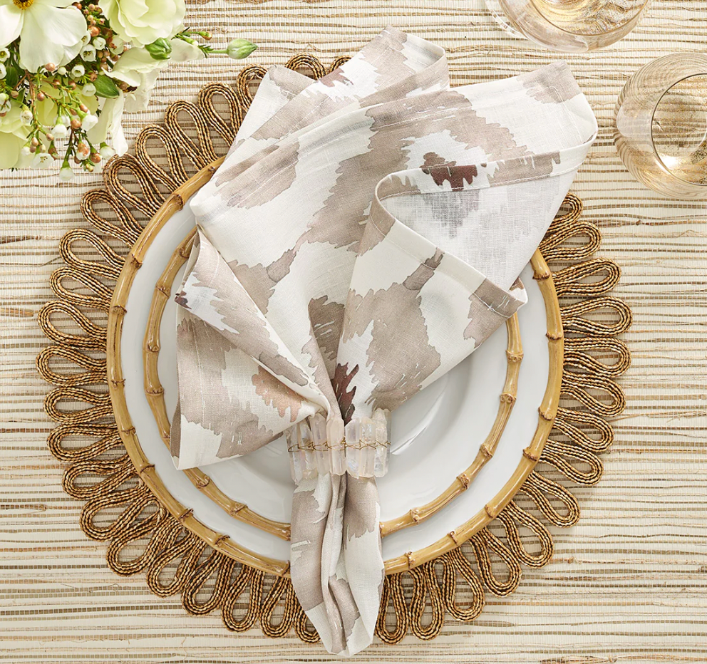 A table setting features a gold placemat, white plate, and a Kim Seybert Watercolor Ikat Napkin Taupe held by a ring, with two glasses. White flowers in the upper left sit atop a striped tablecloth.