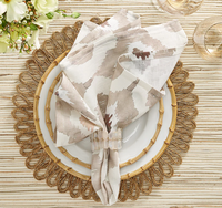 A table setting features a gold placemat, white plate, and a Kim Seybert Watercolor Ikat Napkin Taupe held by a ring, with two glasses. White flowers in the upper left sit atop a striped tablecloth.