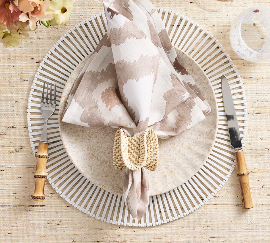 A table setting featuring a round white patterned placemat, beige plate, Kim Seybert Watercolor Ikat Napkin Taupe in a woven ring, bamboo-handled flatware, and a clear glass on a textured surface.