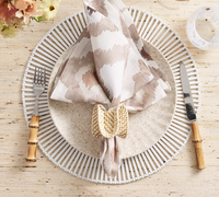 A table setting featuring a round white patterned placemat, beige plate, Kim Seybert Watercolor Ikat Napkin Taupe in a woven ring, bamboo-handled flatware, and a clear glass on a textured surface.