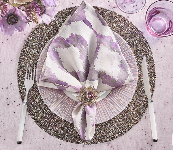 Elegant table setting with a white plate on a beaded placemat, Kim Seybert's Watercolor Ikat Napkin Lilac & Grey Set of 4 in a jeweled ring, silverware, pink glassware, and purple flowers on a pale pink tablecloth.