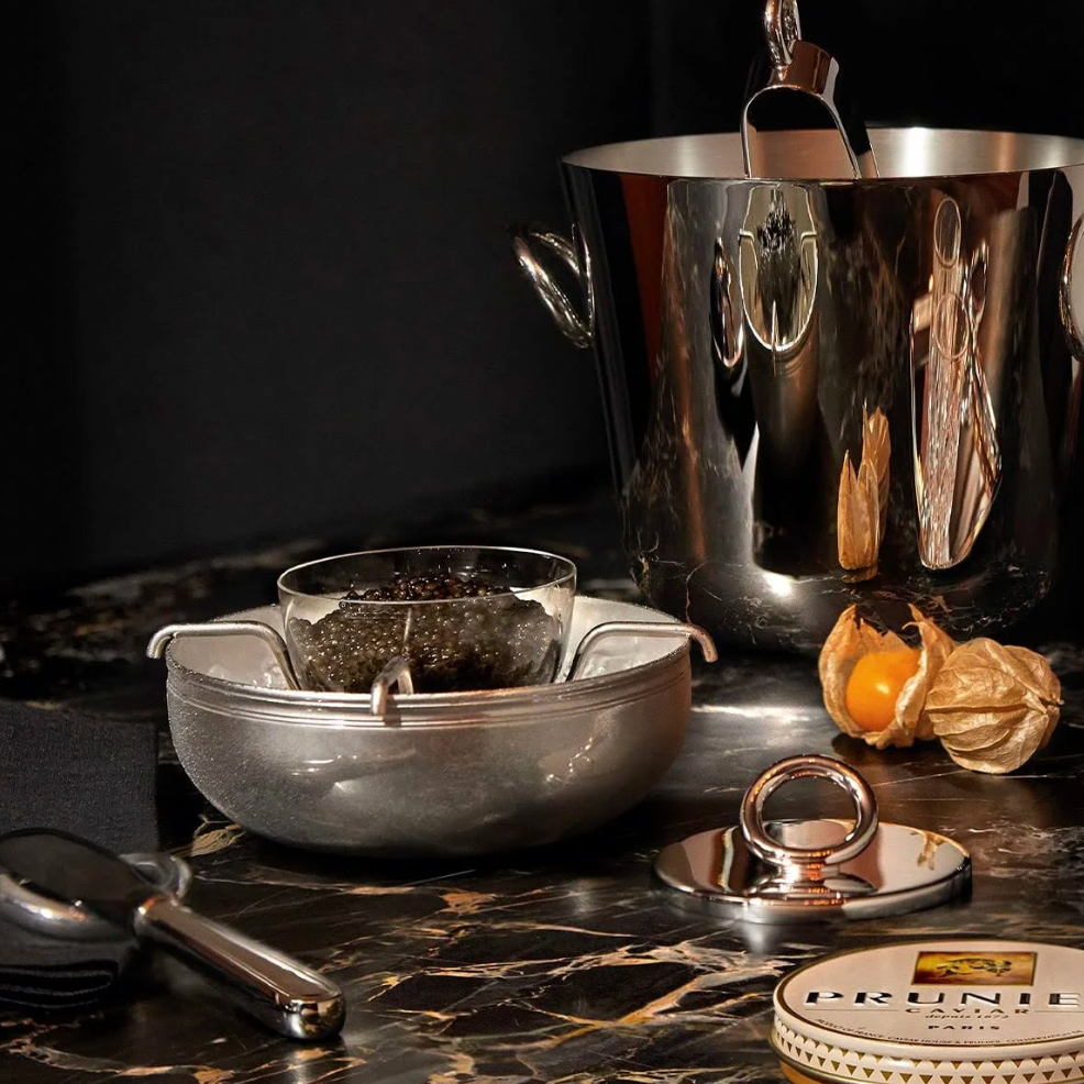 A bowl of black caviar rests on ice in the Christofle Vertigo Silver Plated Caviar Set, accompanied by a silver ice bucket, a Prunier tin, a Vertigo-pattern serving spoon, and a physalis fruit on dark marble.