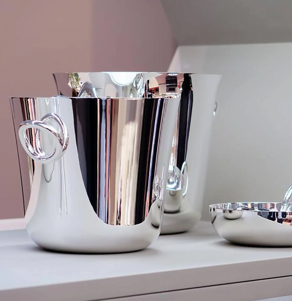 Two Christofle Vertigo Silver Plated Ice Buckets with heart-shaped handles sit beside a matching round bowl on a white surface, their reflective finish enhancing the modern, minimalistic setting.