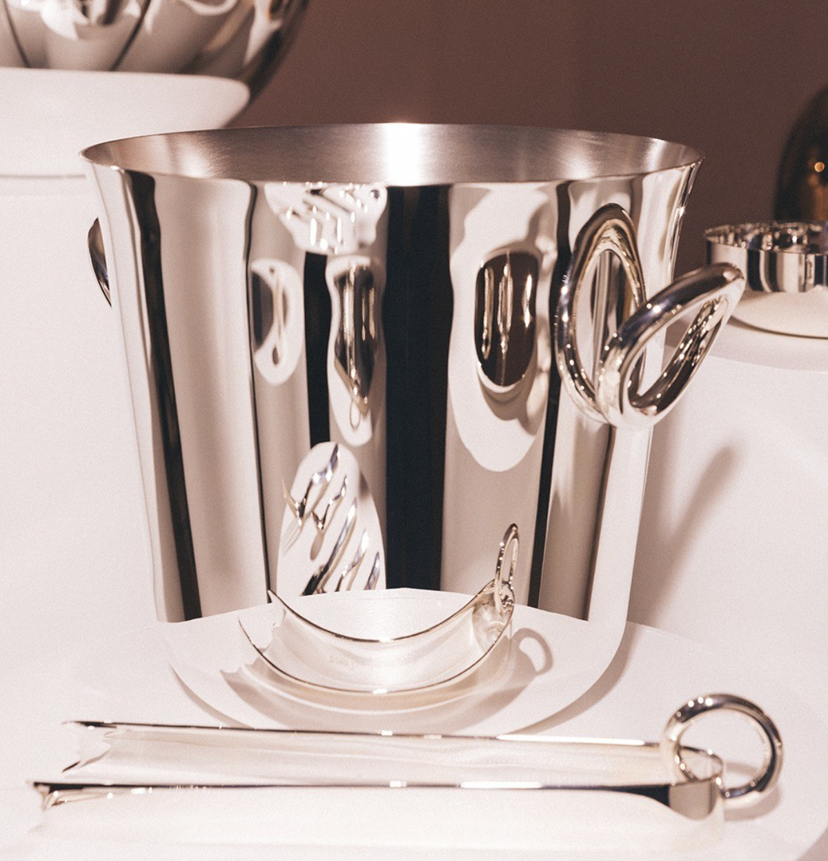 Christofle’s Vertigo Silver Plated Ice Tongs are displayed in the foreground on a white surface, their polished silver finish gleaming under bright light beside a reflective ice bucket with curved handles.