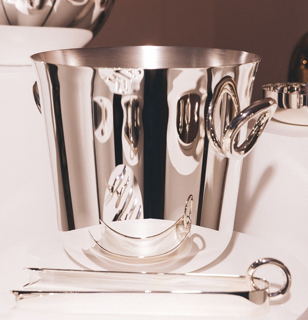 Christofle’s Vertigo Silver Plated Ice Tongs are displayed in the foreground on a white surface, their polished silver finish gleaming under bright light beside a reflective ice bucket with curved handles.