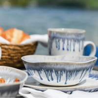 A ceramic bowl, cup, and plate from the Simon Pearce Burlington Dinnerware Pool collection display blue and white drip glaze with glazed edges, arranged outdoors next to a bread basket with water and greenery in the background.