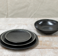 The Tourron Dinnerware Celeste by Jars - Rosenthal sits on a light wooden surface, with three stacked dark gray glazed stoneware plates in varying sizes and a matching bowl placed to the right.