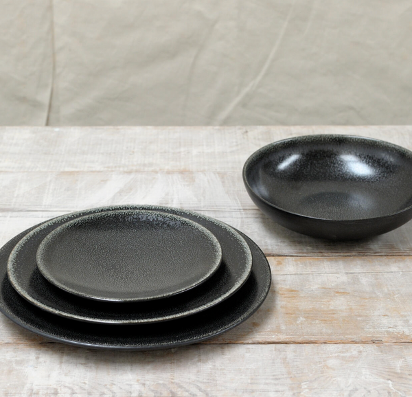 The Tourron Dinnerware Celeste by Jars - Rosenthal sits on a light wooden surface, with three stacked dark gray glazed stoneware plates in varying sizes and a matching bowl placed to the right.