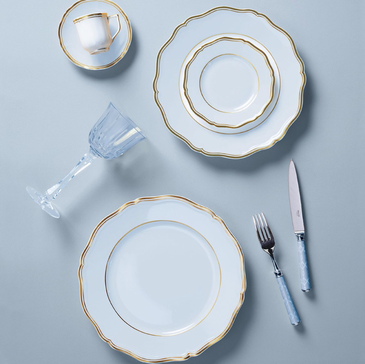 Elegant table setting with Raynaud Mazurka Gold & Blue Grey Dinnerware, including plates, cup and saucer, paired with a crystal wine glass and blue-handled silverware on a light blue surface.