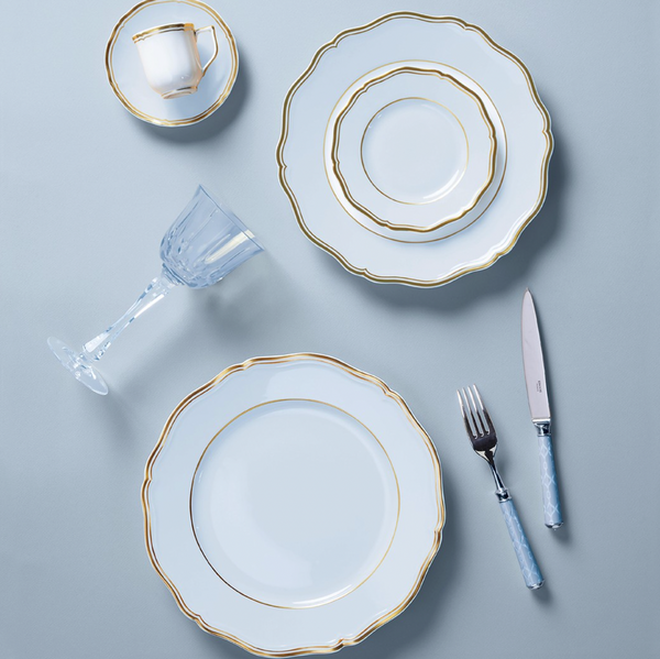 Elegant table setting with Raynaud Mazurka Gold & Blue Grey Dinnerware, including plates, cup and saucer, paired with a crystal wine glass and blue-handled silverware on a light blue surface.