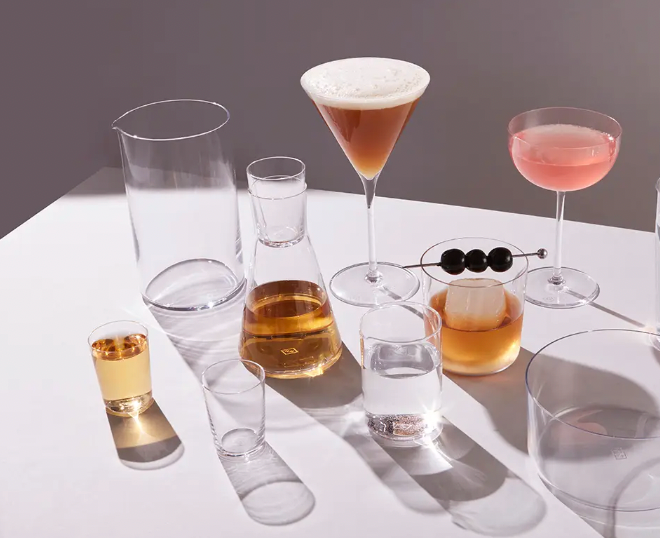 A variety of glassware on a white surface, featuring Richard Brendon’s Cocktail Collection Classic Coupe Glass Set of 2, lead-free crystal cocktail glasses with drinks, a beaker of amber liquid, all bathed in soft lighting with subtle shadows.
