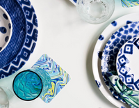 A colorful table setting features blue and white patterned plates, the Vietri Santorini Cereal Bowls Assorted Set of 4, and clear glasses on a white surface with a blue and green marbled coaster under one glass.