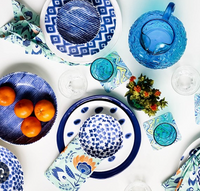 A vibrant table setting features Vietri Santorini Salad Plates Assorted Set of 4 in blue and white patterns, along with bowls, napkins, a blue glass pitcher, water glasses, oranges in a bowl, floral coasters, and a vase of flowers on a white surface.