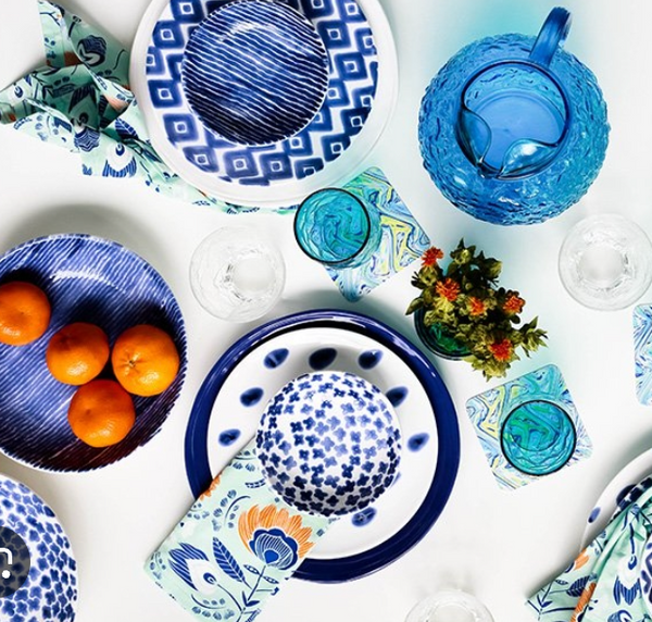 A vibrant table setting features Vietri Santorini Salad Plates Assorted Set of 4 in blue and white patterns, along with bowls, napkins, a blue glass pitcher, water glasses, oranges in a bowl, floral coasters, and a vase of flowers on a white surface.