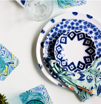 A top view of a table set with Vietri’s Santorini Cereal Bowls Assorted Set of 4, patterned plates, a colorful napkin, glass of water, and blue-green marbled coasters on a white surface.