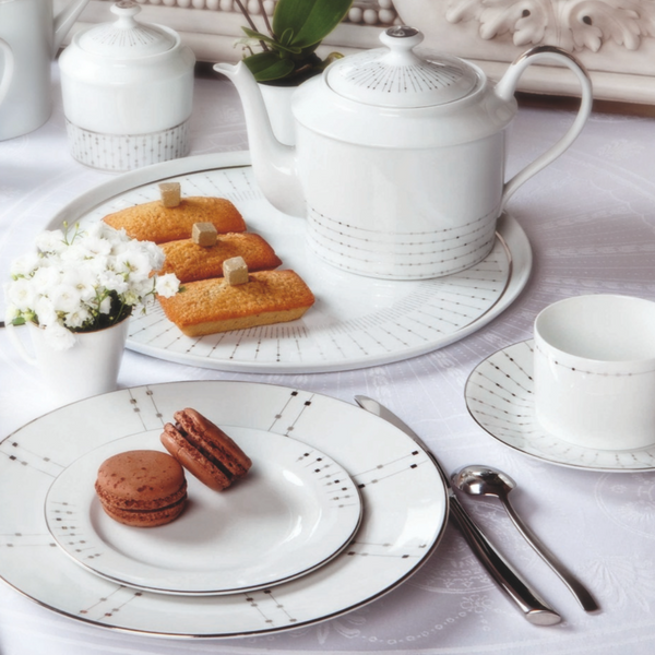 Elegant tea set with Philippe Deshoulieres Carrousel Dinnerware: Limoges porcelain plates, teacup, saucer, teapot with silver accents, two macarons, four cakes on a tray, white flowers, and cutlery on a white tablecloth.