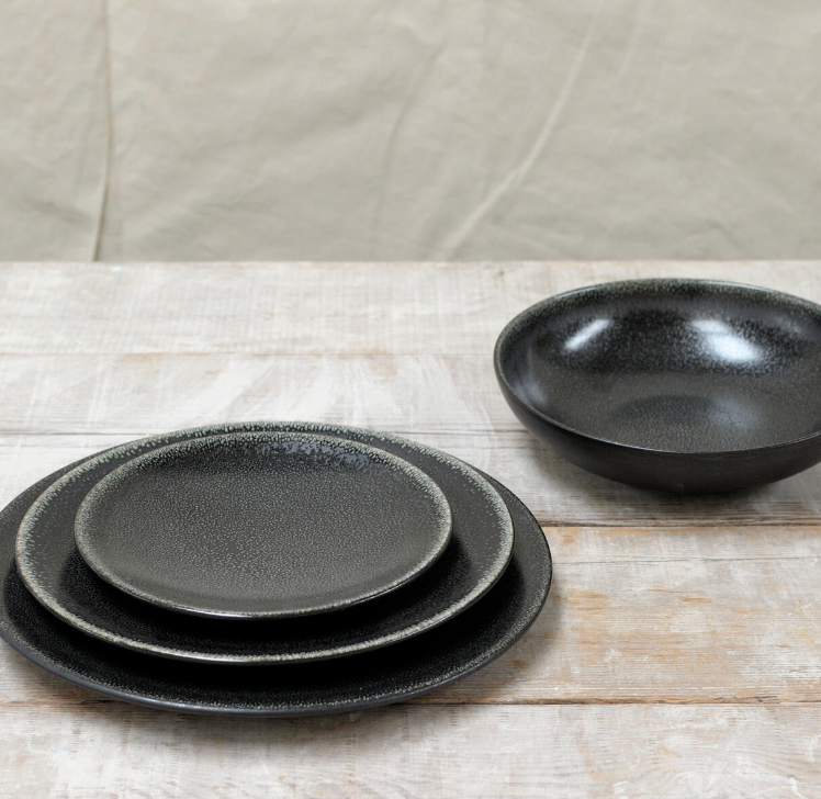 Tourron Dinnerware Celeste by Jars - Rosenthal features three black glazed stoneware plates in varying sizes stacked on rustic wood, with a matching black bowl nearby, set against a neutral wrinkled fabric backdrop.