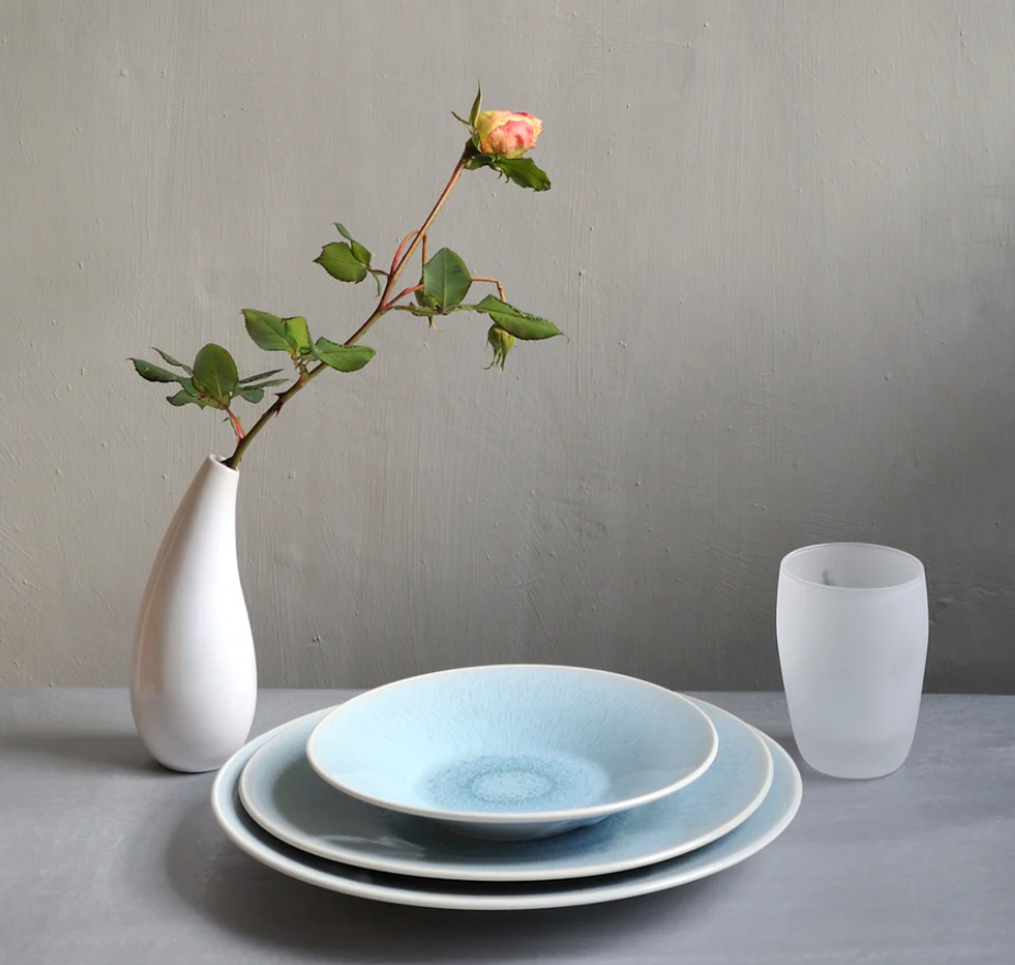 A minimalist table setting features Jars - Rosenthal’s Vuelta Atoll: modern, microwave-safe blue plates stacked with a white vase holding a single pink rose and a frosted glass, all set on a light gray background.