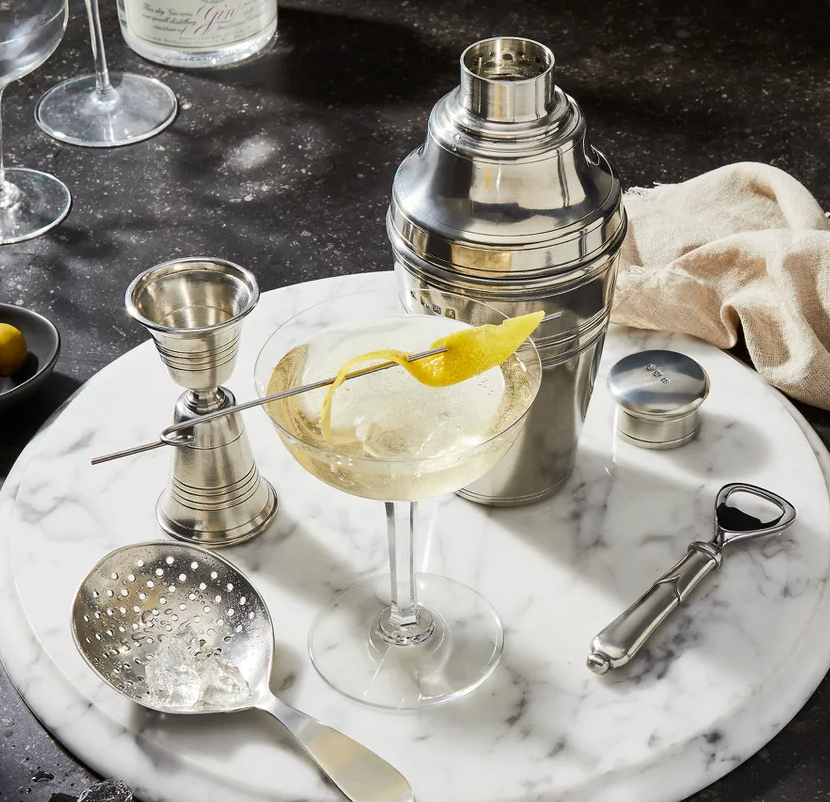 A martini with a lemon twist rests on a marble tray alongside the MATCH Cocktail Shaker, jigger, strainer, peeler, and beige napkin—elegant essentials for any home bar. In the background are a glass of olives and a wine glass.