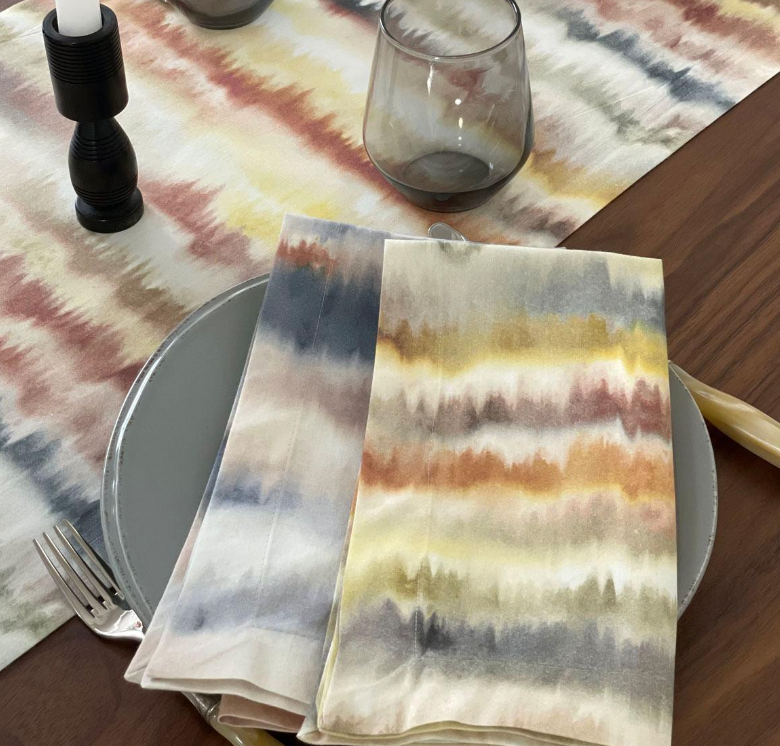 A festive table features Bodrum’s Aria Napkin Desert Set of 4 in yellow, red, and gray, paired with a gray plate, silverware, smoky glass, black candlestick, and a matching machine-washable runner.