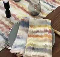 A festive table features Bodrum’s Aria Napkin Desert Set of 4 in yellow, red, and gray, paired with a gray plate, silverware, smoky glass, black candlestick, and a matching machine-washable runner.