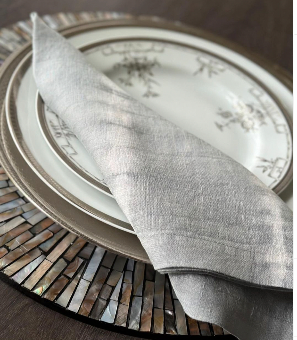 A close-up of an elegant table setting with a white patterned plate, gold charger, and metallic placemat, topped with a neatly folded Hand Dyed Linen Napkin Set of 4 - Pebble by Whigstock Studio.