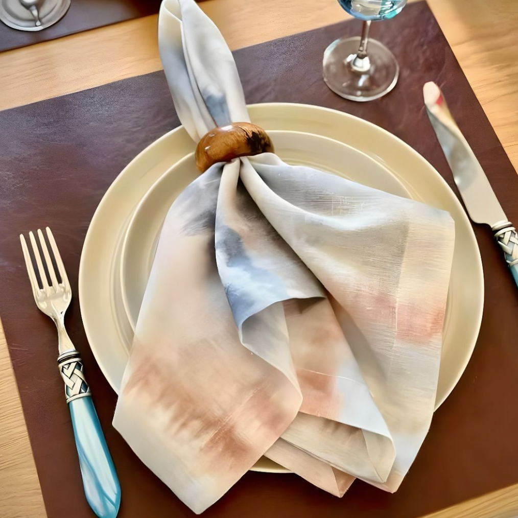 A holiday table setting features a white plate with a Bodrum Aria Napkin Desert (set of 4), held by a wooden ring on a brown placemat, blue-handled fork and knife alongside, and a wine glass.