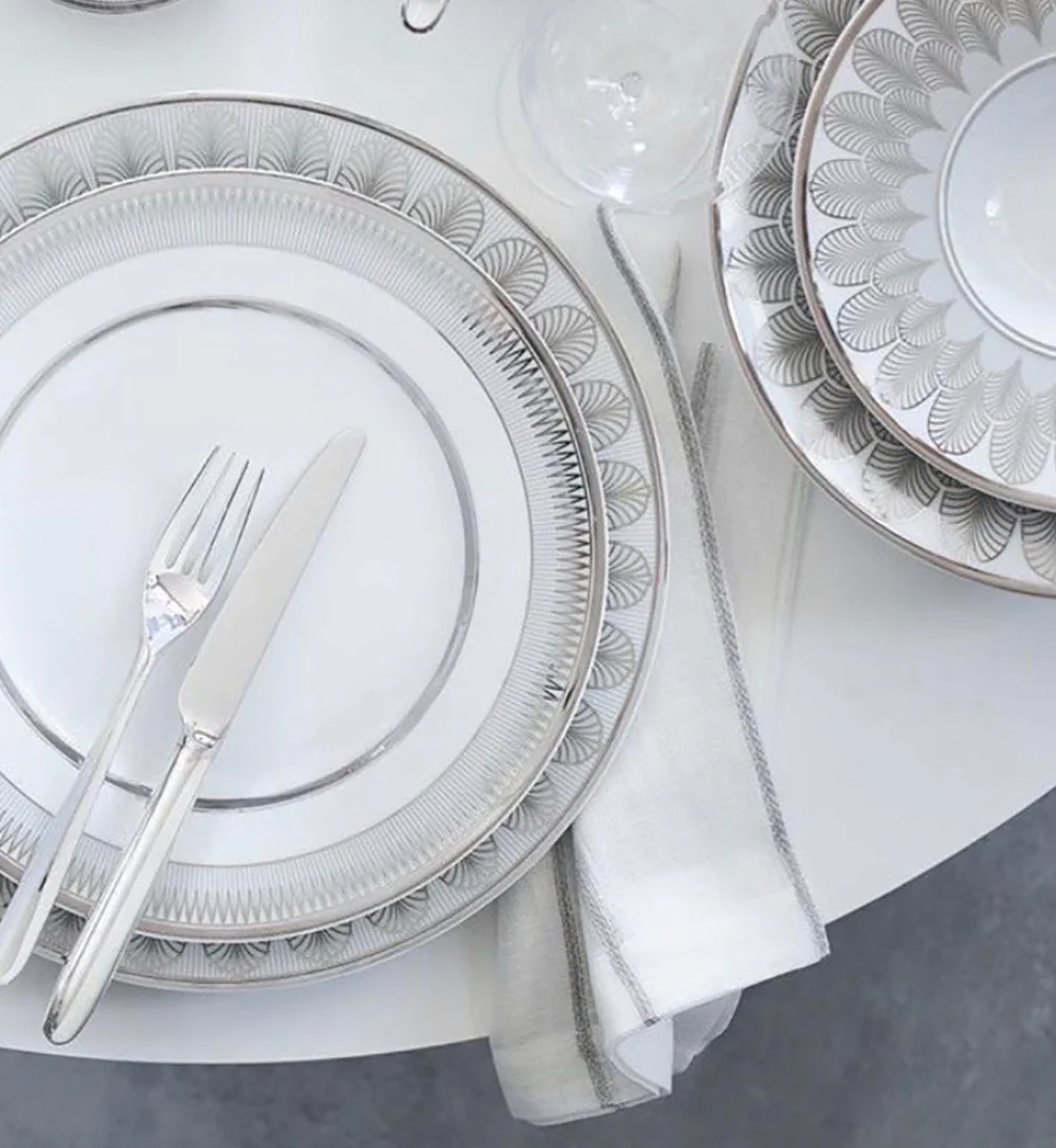 A table set with Christofle Malmaison Imperiale Porcelain Dinnerware Platinum, featuring patterned white and platinum plates, a knife and fork on a dinner plate, stacked bowls, and a folded napkin with platinum trim on a white table.
