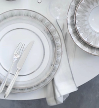 A table set with Christofle Malmaison Imperiale Porcelain Dinnerware Platinum, featuring patterned white and platinum plates, a knife and fork on a dinner plate, stacked bowls, and a folded napkin with platinum trim on a white table.