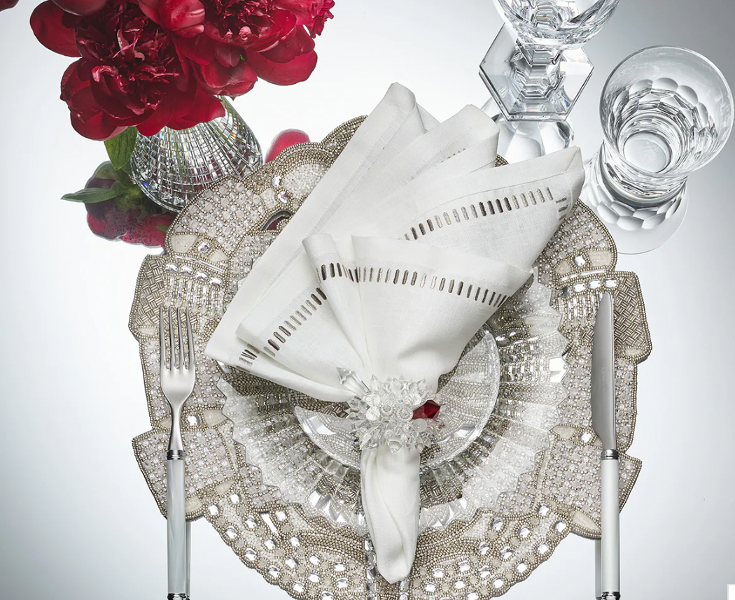 A formal table is set with a silver placemat, clear glass plate, white linen napkin from the Kim Seybert Harmonie Napkin in White, Gray & Silver set, silverware, Baccarat crystal glassware, and a vase of red flowers.