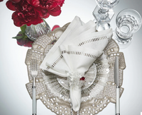 A formal table is set with a silver placemat, clear glass plate, white linen napkin from the Kim Seybert Harmonie Napkin in White, Gray & Silver set, silverware, Baccarat crystal glassware, and a vase of red flowers.