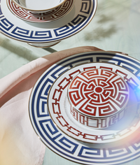 A close-up of Ginori 1735 Labirinto Zaffiro Dinnerware—porcelain plates and a cup with red and blue Greek key patterns—on a glass table with a pale pink napkin, sunlight casting a soft lens flare.