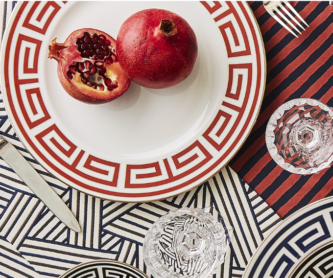 A table setting showcases a Ginori 1735 Labirinto Dinnerware Nero porcelain plate with geometric design, holding whole and halved pomegranates on a patterned tablecloth, accompanied by crystal glasses, a knife, and a fork.
