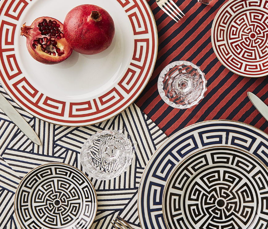 A table is set with Ginori 1735 Labirinto Zaffiro dinnerware featuring red and black Greek key patterns, glass cups, silverware, and striped geometric tablecloths. A whole and halved pomegranate rest on one of the plates.