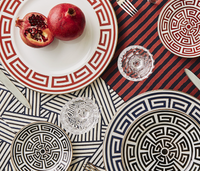 A table is set with Ginori 1735 Labirinto Zaffiro dinnerware featuring red and black Greek key patterns, glass cups, silverware, and striped geometric tablecloths. A whole and halved pomegranate rest on one of the plates.