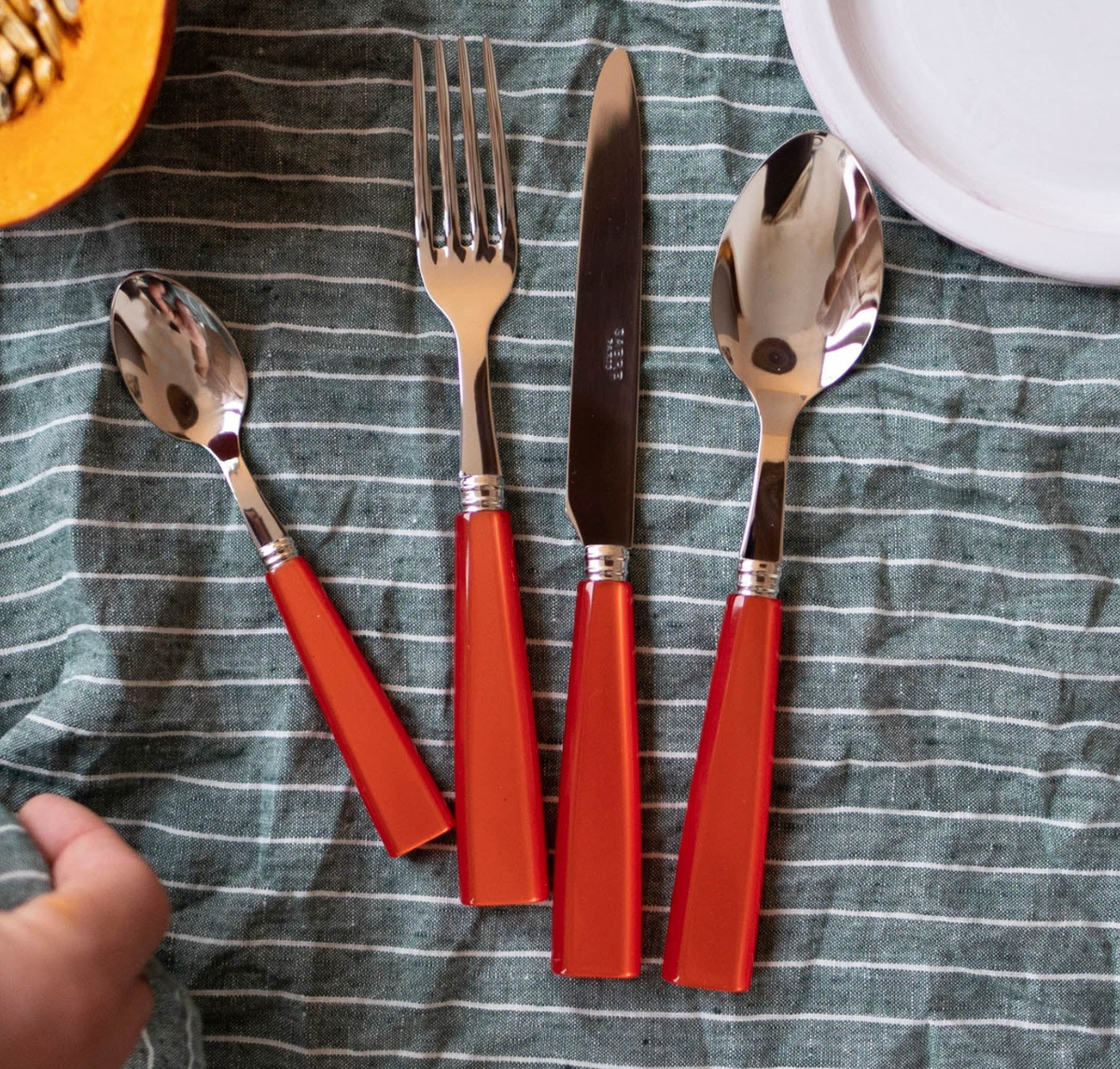 The Sabre Icone Flatware 5 Piece Setting, featuring shiny 18/10 stainless steel tops and red handles, is arranged on a green and white striped cloth beside a partial white plate and a hand holding cut orange squash.