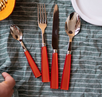The Sabre Icone Flatware 5 Piece Setting, featuring shiny 18/10 stainless steel tops and red handles, is arranged on a green and white striped cloth beside a partial white plate and a hand holding cut orange squash.