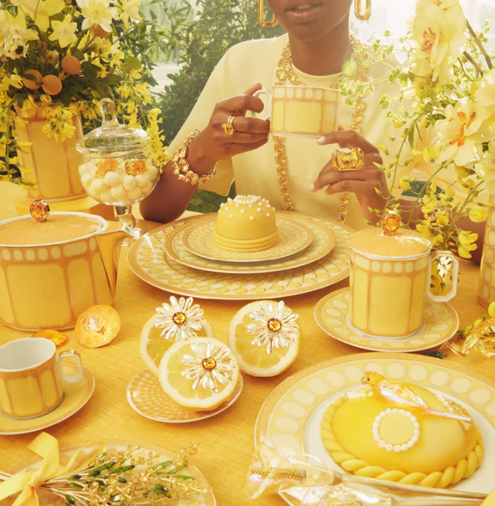 A person sits at a table adorned with Swarovski x Rosenthal Signum Jonquil Yellow Mugs, surrounded by yellow flowers, gold jewelry, and floral-themed desserts for an elegant, radiant atmosphere.