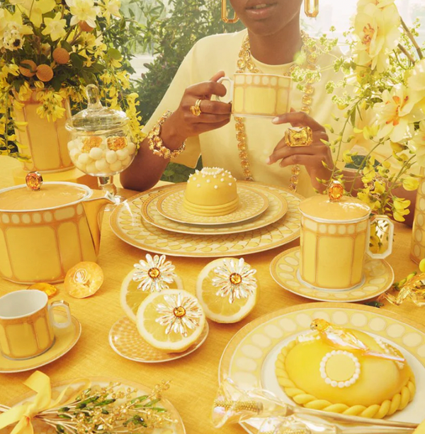 A person sits at a table adorned with Swarovski x Rosenthal Signum Jonquil Yellow Mugs, surrounded by yellow flowers, gold jewelry, and floral-themed desserts for an elegant, radiant atmosphere.