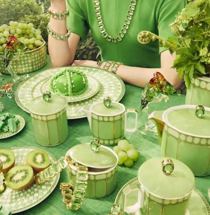 A woman in green attire sits at a table with Swarovski x Rosenthal Signum Fern Green Mugs, surrounded by green-themed plates, fruits, herbs, and gold accents—an ideal luxury gift box scene.