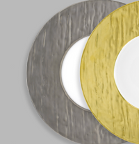 Two overlapping Makassar Metallic Charger plates by Raynaud, one gold and one silver, rest on a gray background. Each plate has a textured, uneven surface with a white center, and only part of each is visible.