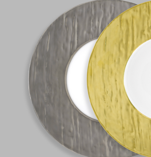 Two overlapping Makassar Metallic Charger plates by Raynaud, one gold and one silver, rest on a gray background. Each plate has a textured, uneven surface with a white center, and only part of each is visible.