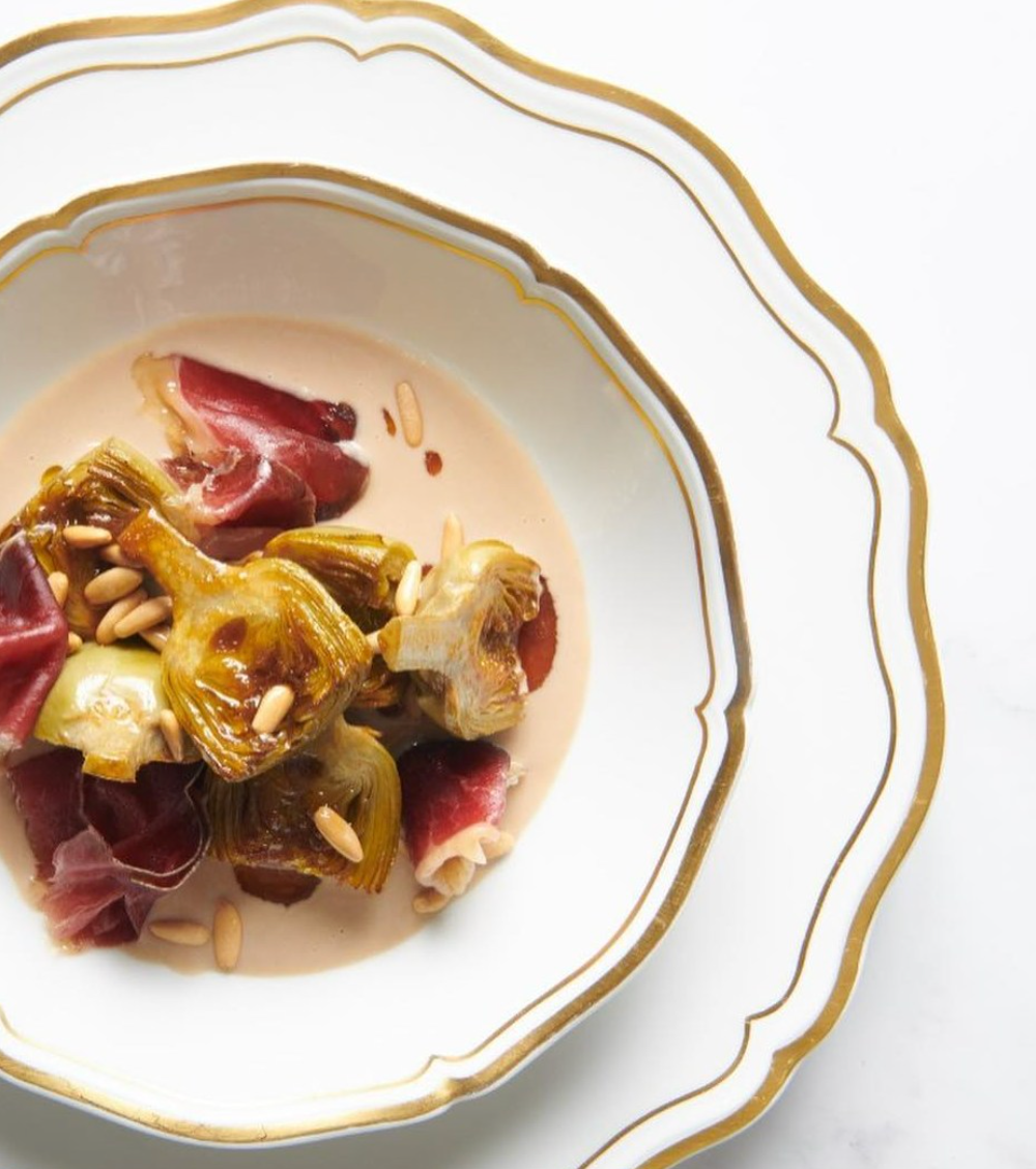 A gourmet dish of caramelized artichokes, cured meat, pine nuts, and creamy sauce is artfully presented on a white Polka Gold Dinnerware plate by Raynaud.